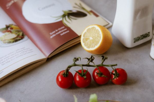 Flat lay of cooking ingredients including tomatoes and a lemon with a cookbook.