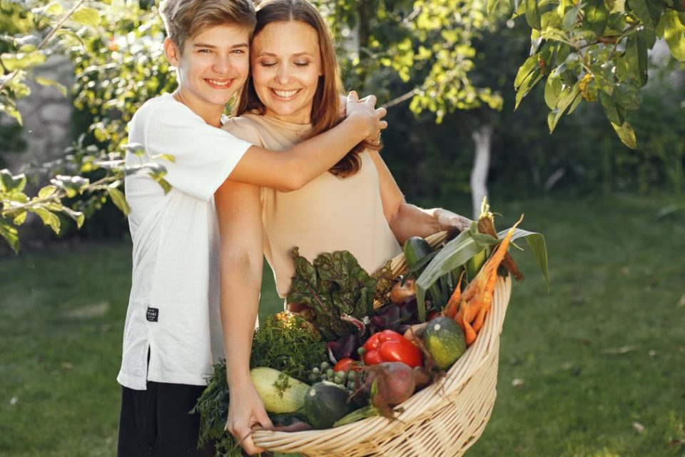 Mother and son smiling outdoors with a basket full of fresh vegetables, embracing in a sunny garden.